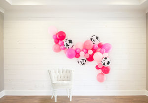 Various pinks ranging from soft to neon plus cow print balloons made into a balloon garland that is hanging on a white shiplap wall with a chair in front of it.