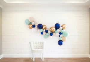 A nautical themed balloon garland is draped across a white wood wall with a cloth white chair in front. The garland consists of various 5 inch and 11 inch balloons in the colors of pearl white, tan, jade, navy, and chrome gold.