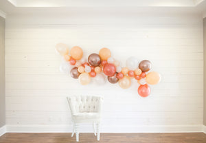 A bridal themed balloon garland is draped across a white wood wall with a white cloth chair in front.