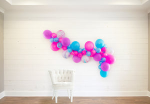 The neon themed garland is draped along a white wood wall with a white cloth chair sitting in front to help give a sense of the garlands size and length.