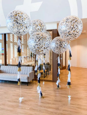 Five 3ft clear balloon stuffed with tissue confetti and tissue tassel string in the colors white, black, and gold sitting in an office.