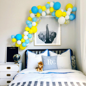 A balloon garland in the colors white, blue, and yellow hangs above a black bed. Above the bed and under the garland is a black and white elephant photo. On the bed rests a stuffed animal lion and there are white and blue bed covers.