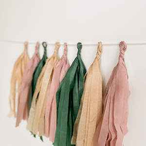 A side view of eight hand rolled tassles in the colors tan, mauve, and forest green on a white background.