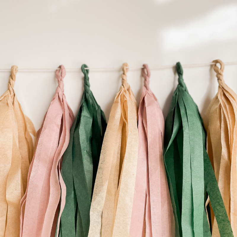 A photo of 7 hand rolled tassles in the colors tan, mauve, and forest green on a white background.