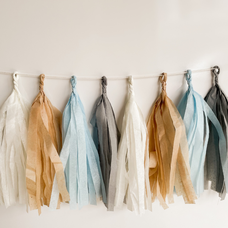 Eight hand rolled tissue tassel garlands hung from a white string on a white wall. The tassel colors are cream, tan, light blue, and gray. 