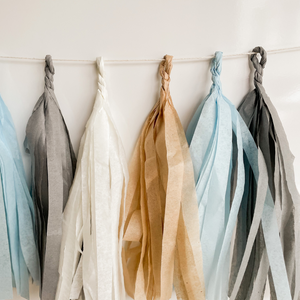 Eight hand rolled tissue tassel garlands hung from a white string on a white wall. The tassel colors are cream, tan, light blue, and gray. 