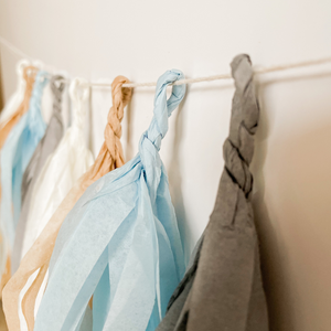 Eight hand rolled tissue tassel garlands hung from a white string on a white wall. The tassel colors are cream, tan, light blue, and gray. 