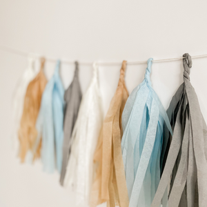 Eight hand rolled tissue tassel garlands hung from a white string on a white wall. The tassel colors are cream, tan, light blue, and gray. 