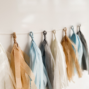 Eight hand rolled tissue tassel garlands hung from a white string on a white wall. The tassel colors are cream, tan, light blue, and gray. 