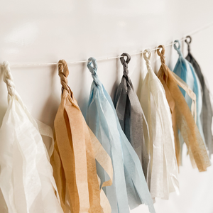 Eight hand rolled tissue tassel garlands hung from a white string on a white wall. The tassel colors are cream, tan, light blue, and gray. 