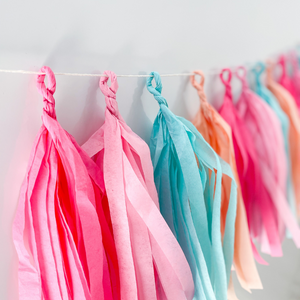 Close up of 12 hand rolled tissue tassels hanging from a white string used as party decor against a white wall.