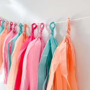 Side view of 12 hand rolled tissue tassels hanging from a white string used as party decor against a white wall.