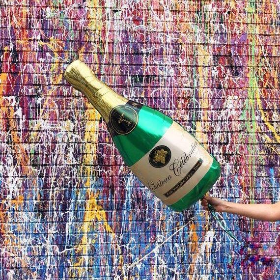 A champagne mylar balloon is being held in front of a crazy streak painted brick wall. The champagne bottle has a gold colored neck, and green body, and a tan colored label around it with dark lettering.