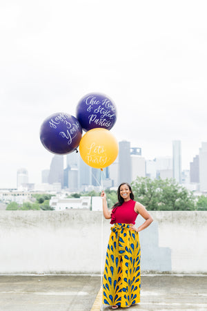 Woman standing in a parking garage is holding three jumbo 36 inch balloons; two purple and one yellow.