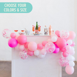 A balloon garland sits draped across the front and side of a beverage cart that has a bottle and multiple glasses sitting on it. The garland is made of pink, rose, white, neon magenta, and coordinating confetti balloons.
