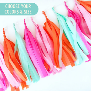 A arial shot of a hot pink, pink, orange, and mint tissue tassel garland is taken of tassels strung on twine and laying on a white table. The tassels are approximately nine inches long.