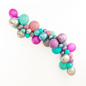 Balloon garland arch in the colors chrome silver, chrome purple, chrome blue, caribbean blue, aquamarine, spring lilac, gray, silver, soft plum, and neon purple is displayed on a white wall at a celebration.