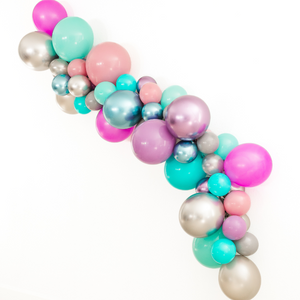 A balloon garland with the following colors: chrome silver, chrome purple, chrome blue, caribbean blue, aquamarine, spring lilac, gray, silver, soft plum, and neon purple is hung on a white wall at a party.