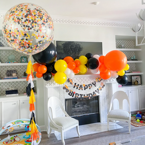 Clear jumbo confetti filled balloon with matching tassel tail and a garland above a chimney. The party colors are white, black, orange, and yellow.