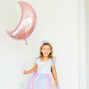 Little girl wearing a fairy costume is sticking her tongue out and holding a pink crescent moon jumbo balloon.