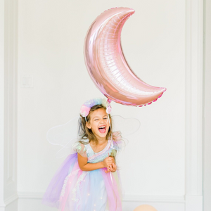 Little girl wearing a fairy costume is smiling and holding a pink crescent moon jumbo balloon.