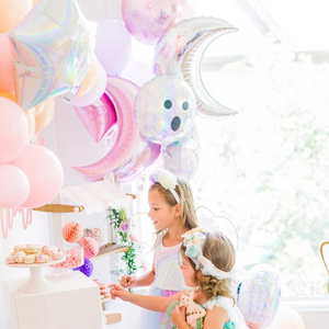 Two little girls dressed in fairy costumes are looking at a table full of treats. Behind them there are balloons in moon and star shapes.