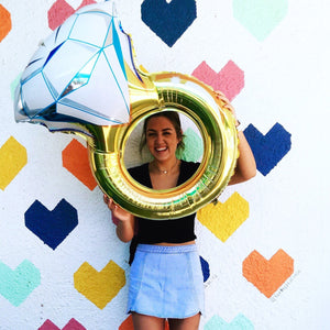Woman holding a jumbo diamond ring balloon in front of a wall with hearts painted on.
