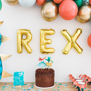 Gold balloon letters spell REX above a chocolate cake at a three year olds dinosaur themed birthday party.