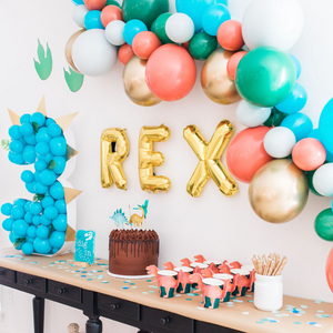 A balloon garland hangs on the wall in the colors tropical teal, green, chrome gold, green blend, and coral blend. The birthday theme is dinosaurs; there is a three made out of blue balloons and gold letter balloons spell REX sits next to it.
