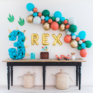 A dinosaur themed birthday party for a three year old. There is an 8ft balloon garland with the colors tropical teal, green, chrome gold, green blend, and coral blend. Below the balloon garland are three gold letter balloons spelling REX and a number 3 made out of small blue balloons. A black wood table sits below the balloon garland with a chocolate cake, dinosaur toppers and cups, and hand cut 1" blue confetti.