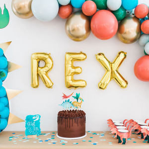 A balloon garland with the colors tropical teal, green, chrome gold, green blend, and coral blend hang on a wall. Below the balloon garland are gold letter balloons spelling REX and a chocolate cake sits below it with custom tissue confetti around the cake.