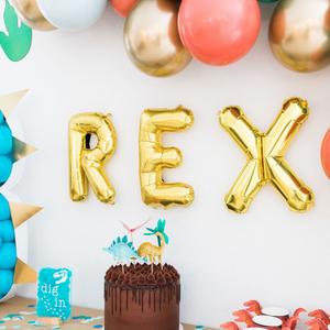 A close up of a dinosaur themed birthday party. Gold letter balloons spell REX and above it is a balloon garland.