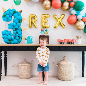 A little boy stands in front of a dinosaur themed birthday party. A black wood table behind him has a giant number 3 on top, gold letter balloons spelling REX, a long balloon garland, and a chocolate cake.