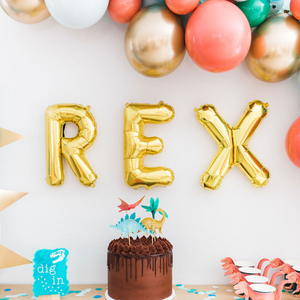 Dinosaur themed three year olds birthday party. There are gold letter balloons spelling REX on the wall and below it sits a chocolate icing cake with dino toppers.