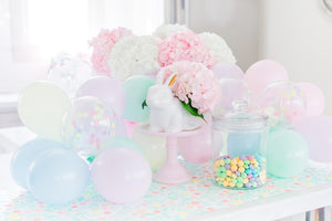 A white bunny sits on a pastel pink cake stand surrounded by pastel colored mini balloons.