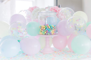 A clear jar with pastel colored MandM chocolates sits on a pastel pink cake stand surrounded by pastel colored mini balloons.