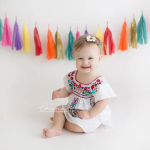 A little girl is sitting at the center and wearing a white fiesta style dress. Behind her is a banner of hand rolled tassels.