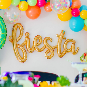 A gold cursive balloon that says fiesta is hung on a wall underneath a fiesta themed balloon garland.