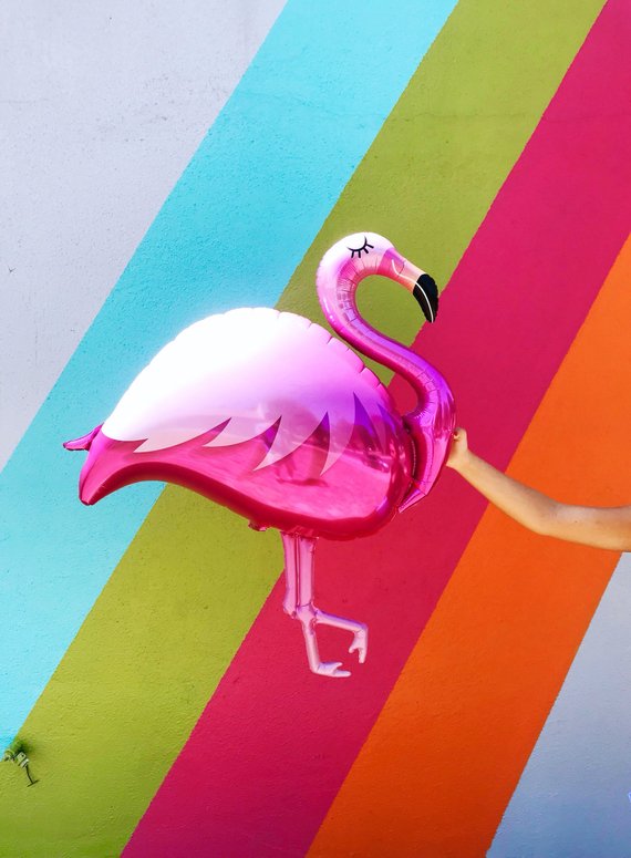Bright pink flamingo balloon being held by a womans hand in front of a brightly colored wall.