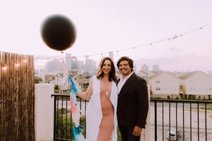 Couple standing on a balcony holding a black jumbo balloon filled with either pink or blue confetti.