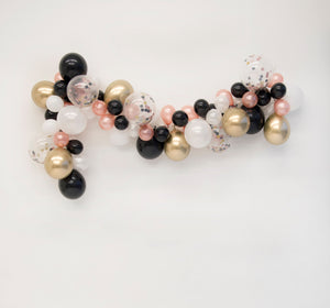 A pretty New Years Eve balloon garland is draped across a white wall made up of white, black, rose gold, chrome gold, and matching confetti balloons.