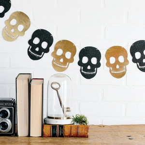 A halloween black and gold skull banner hangs on the wall. Below the banner are three books and a gold key inside a cloche.