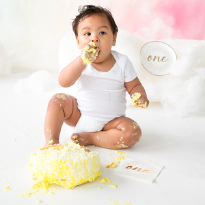 Little boy eating a yellow cake with a white napkin with gold foil cursive ONE written on it next to him and a matching round plate.