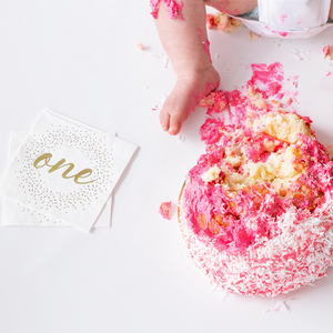 White napkins with gold foil cursive ONE written on it and gold confetti dots next to a pink smash cake and a baby girls foot.