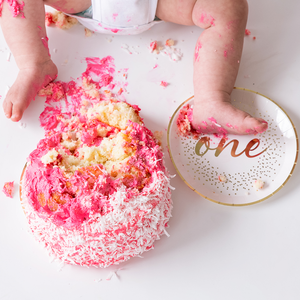 Two baby feet peek from the top of the photo, one foot is near a pink frosting birthday cake and the other is on top of a white and gold paper plate that says ONE.