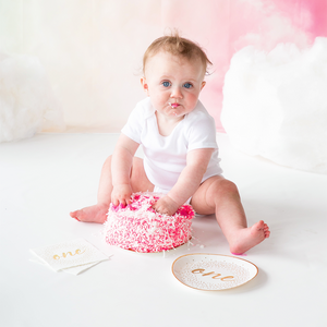Little baby smashing a pink frosting cake with white and gold napkins and plates next to the cake.