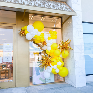A balloon garland hangs on the glass window of a Kendra Scott jewelry store. The balloons are white and yellow with gold star balloons.