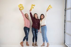 Three girls stand in a white room each holding a gold letter balloon in their hands as high as they can facing the camera and smiling. The letter balloons are BFF.