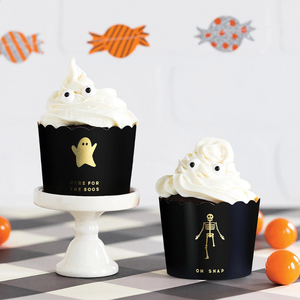 Halloween party decor with two black food cups. One reads HERE FOR THE BOOS in gold foil letters with a gold ghost above it. The other food cup reads OH SNAP with a gold foil skeleton missing one leg.