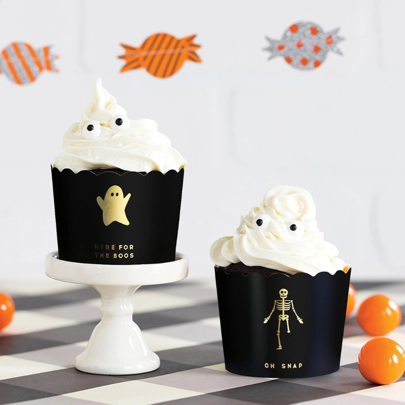 Halloween party decor with two black food cups. One reads HERE FOR THE BOOS in gold foil letters with a gold ghost above it. The other food cup reads OH SNAP with a gold foil skeleton missing one leg.